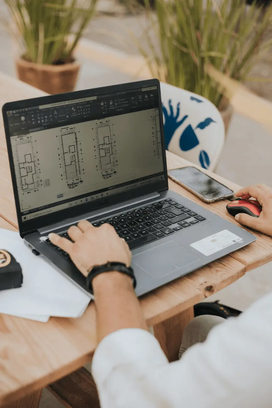 A CAD drafter is working on a laptop with architectural drawings open in AutoCAD. The person is using a mouse and keyboard while sitting at a wooden table, with a tape measure, a smartphone, and plants in the background.