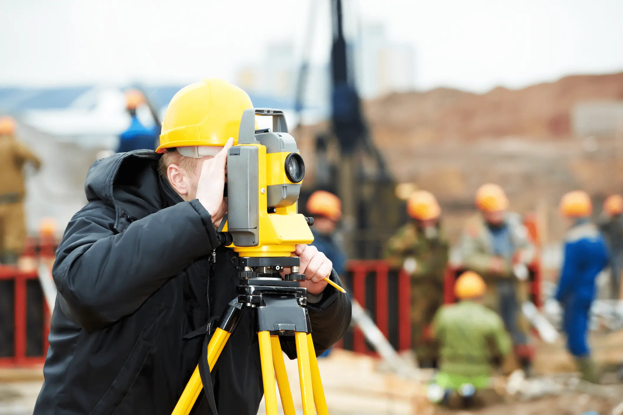 A surveyor is wearing a yellow hard hat and a black jacket while using a surveying instrument on a construction site. In the background, construction workers are working in hard hats and work clothes.