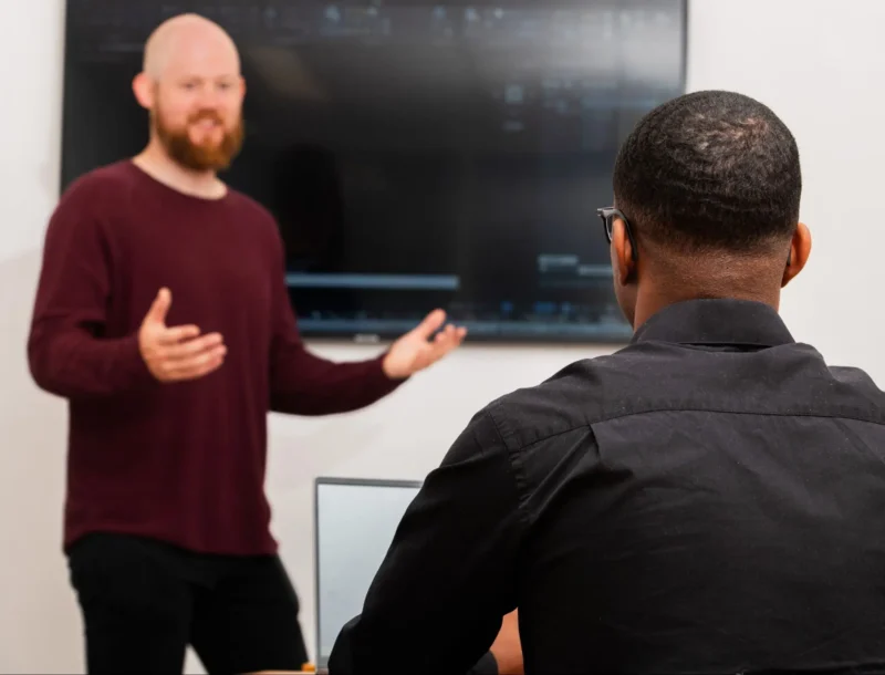 Man giving a presentation to colleague in front of a large screen in an office setting.