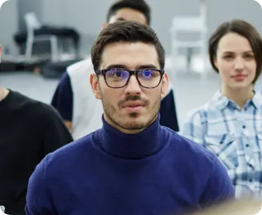 Man in glasses and blue sweater attentively listening in a classroom setting with peers in the background.