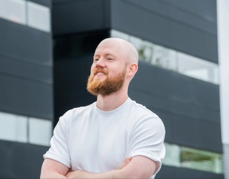 Smiling man with beard outdoors wearing white shirt; modern glass building in background.