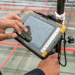 Een maatvoerder gebruikt een Topcon FC-5000 tablet om metingen te verrichten op een bouwplaats. De robuuste tablet is bevestigd aan een meetinstrument en toont een digitale bouwtekening. Op de achtergrond zijn stalen wapeningsnetten en rode meetlijnen zichtbaar.