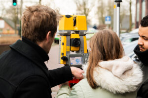Drie mensen die samenwerken met een gele total station op een trottoir, waarbij ze punten aan het inmeten zijn en de meetresultaten bekijken op het scherm van de total station.