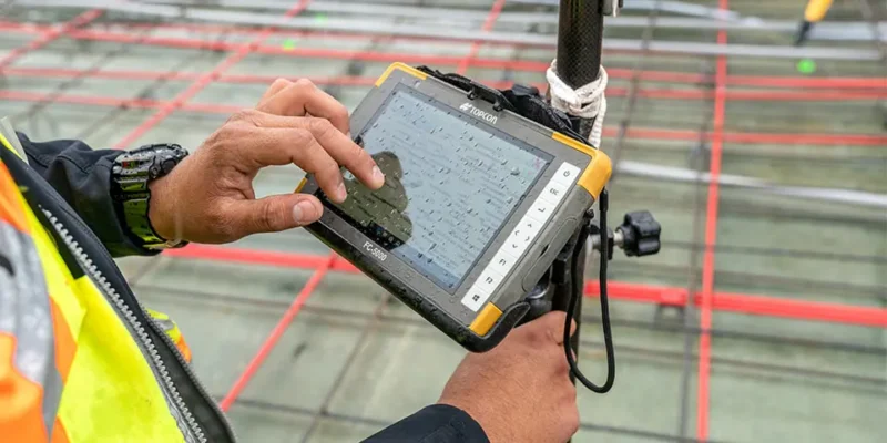 Een maatvoerder gebruikt een Topcon FC-5000 tablet om metingen te verrichten op een bouwplaats. De robuuste tablet is bevestigd aan een meetinstrument en toont een digitale bouwtekening. Op de achtergrond zijn stalen wapeningsnetten en rode meetlijnen zichtbaar.