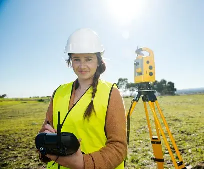 Landmeetkundige met helm en hesje bedient dronecontroller naast total station tijdens veldwerk in open gebied