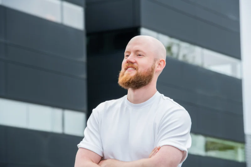Professioneel portret van lachende kale man met rode baard in witte trui voor modern kantoorgebouw